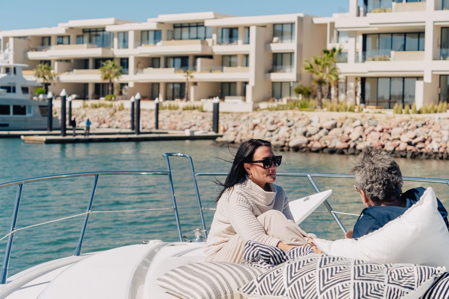 People relaxing on a boat deck while sailing along a waterfront with modern apartment buildings in the background.