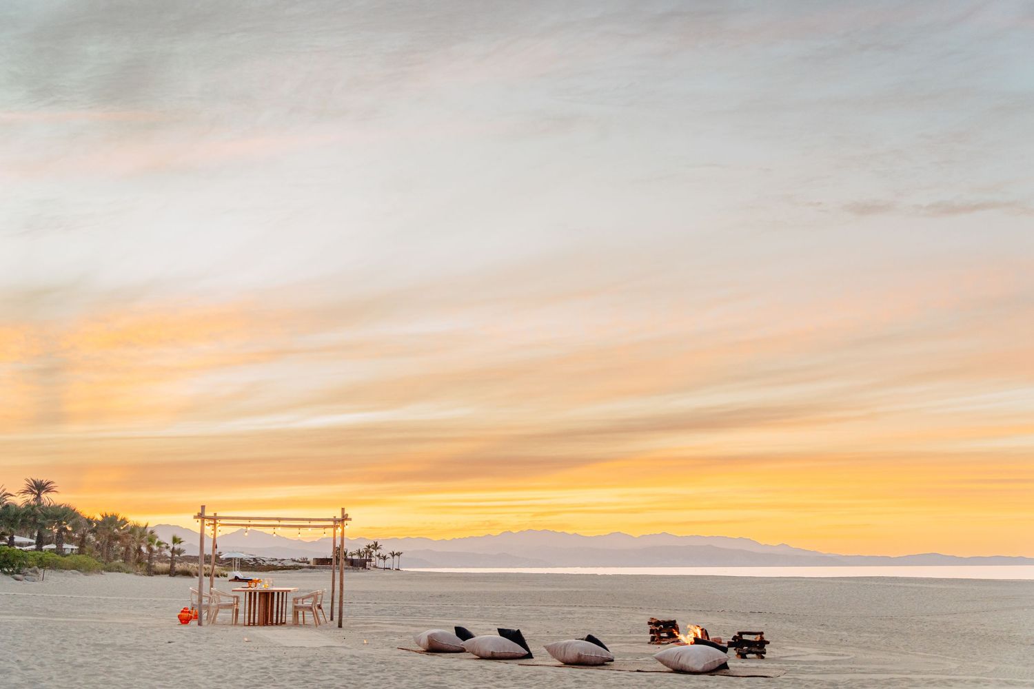 A stunning orange and pink sunset illuminates the sky over a tranquil beach with silhouetted palm trees and chairs.