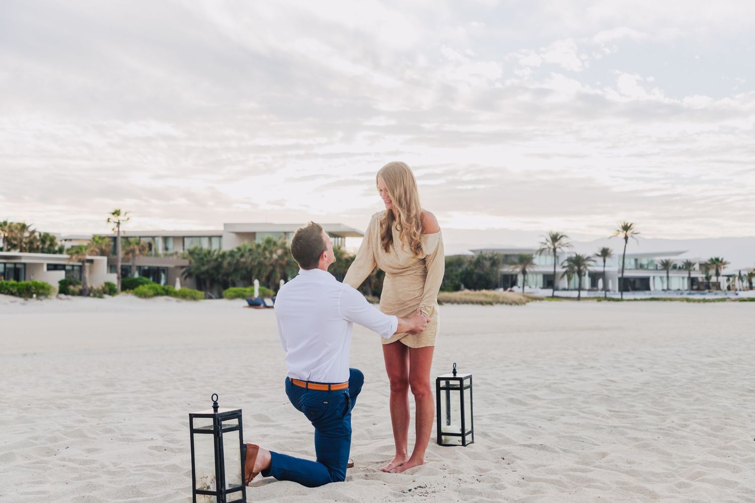 A proposal scene unfolds on sandy beach with decorative wooden arch and lanterns.