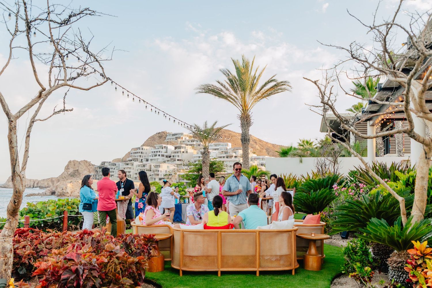 A panoramic view of an outdoor deck with palm trees and people gathered for a beachside event at sunset.
