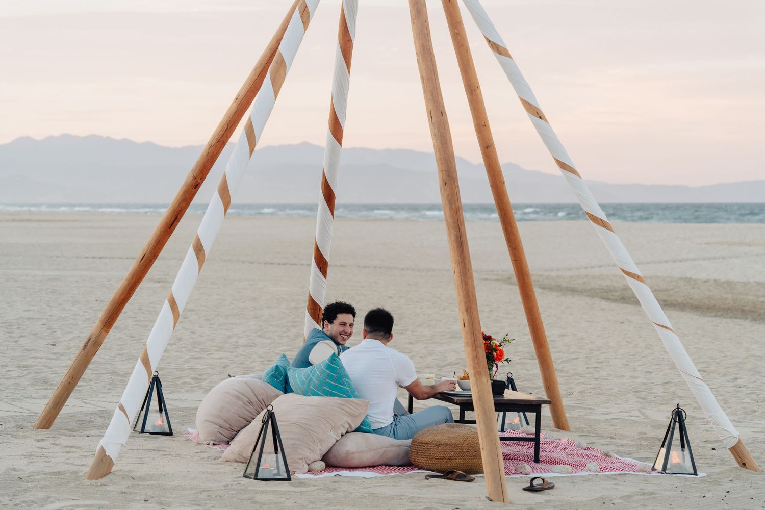 Romantic beach proposal setup with wooden teepee frame and lanterns at sunset.