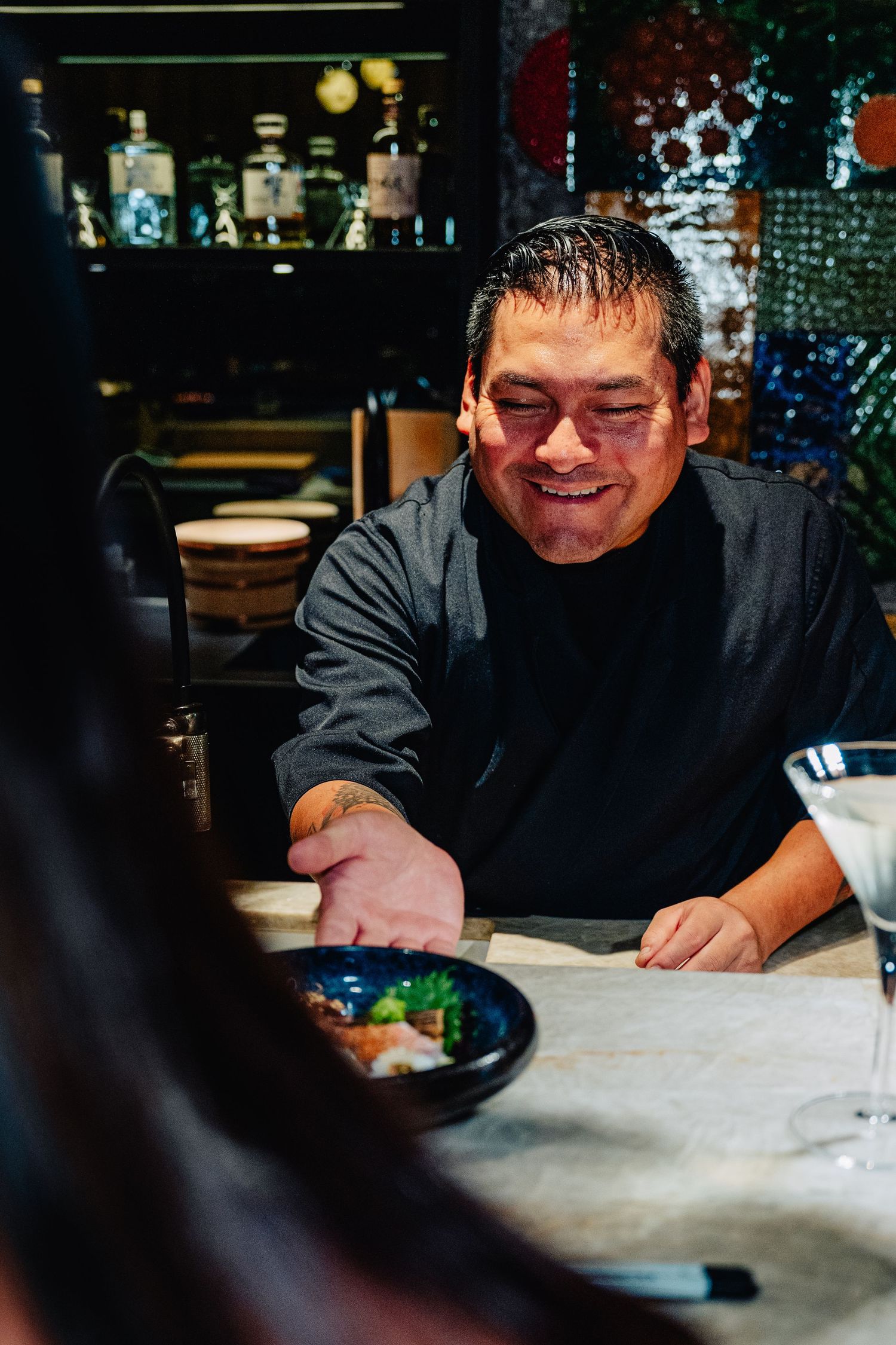 Chef in black uniform focused on preparing dishes at a dimly lit restaurant counter.