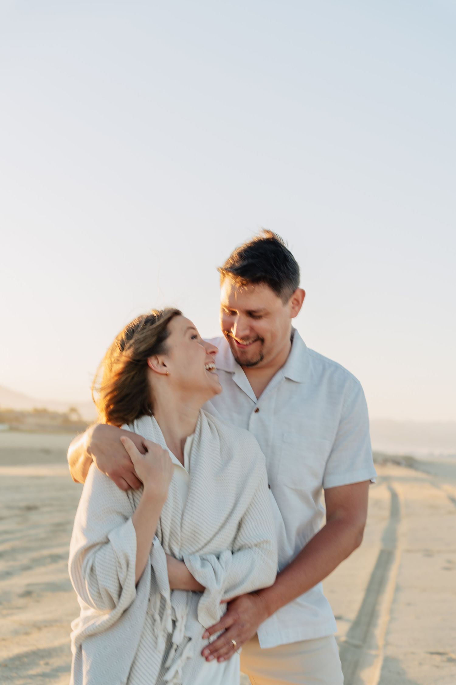 Romantic couple in white outfits embrace and laugh together on a sandy beach during golden hour sunset.