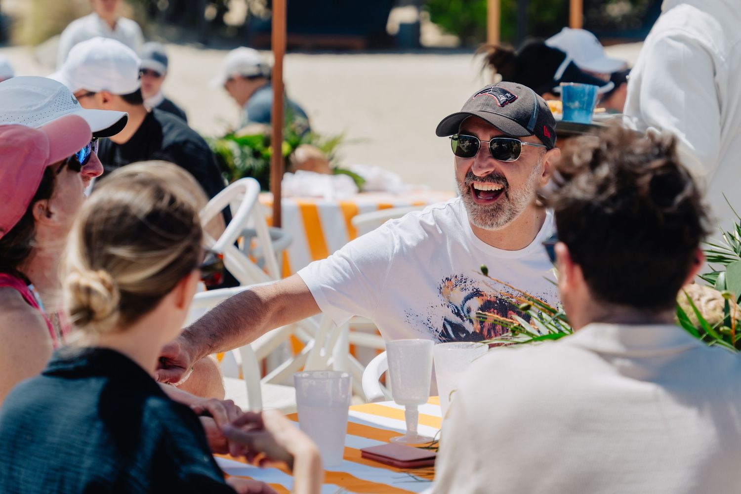 A group of people enjoying drinks and conversation at an outdoor table on a sunny day.