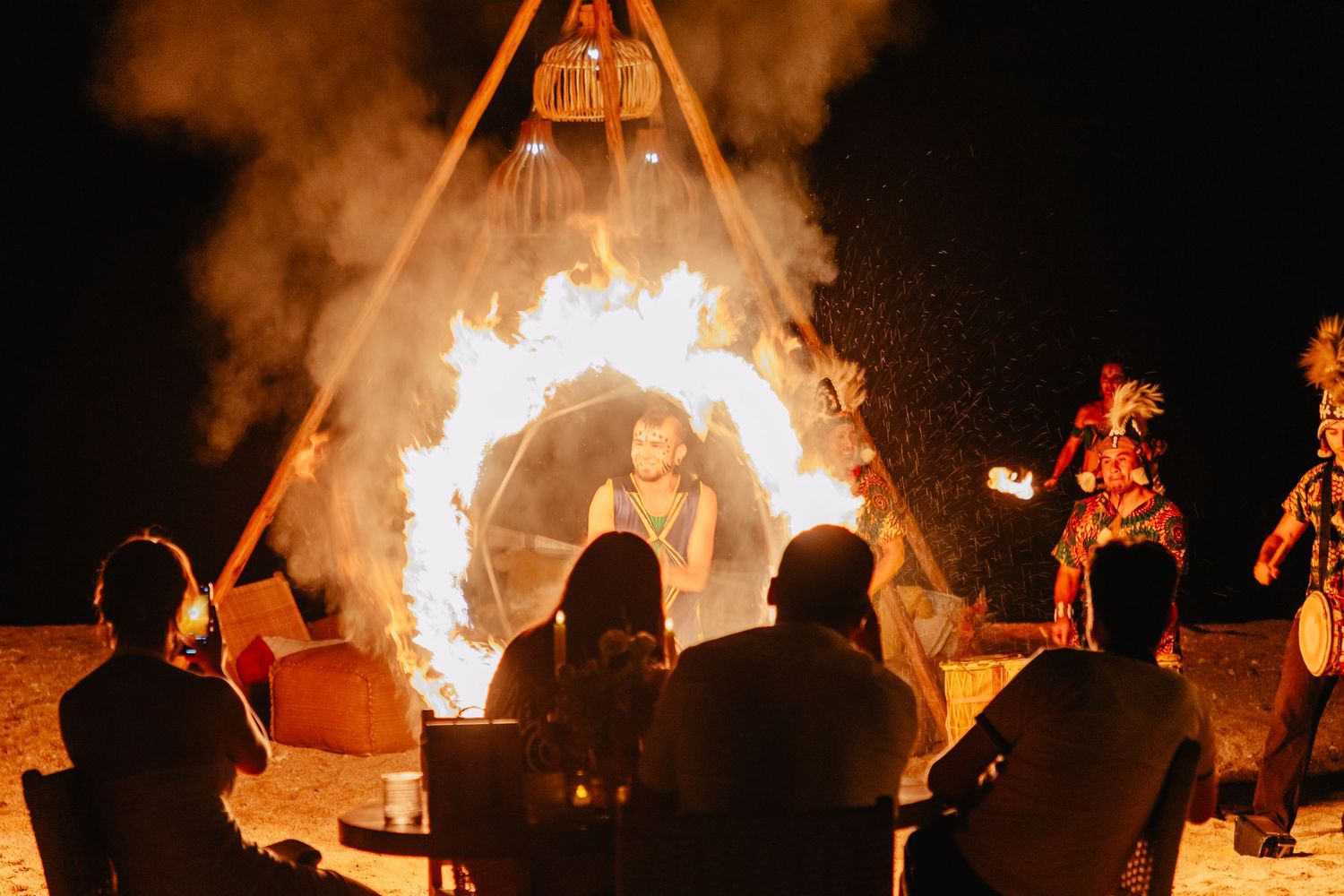 Silhouettes of people gathered around a large bonfire at night with smoke rising into the dark sky.
