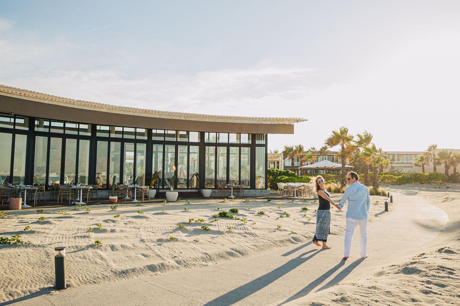 Couple strolling along sandy path near modern glass building during golden hour.