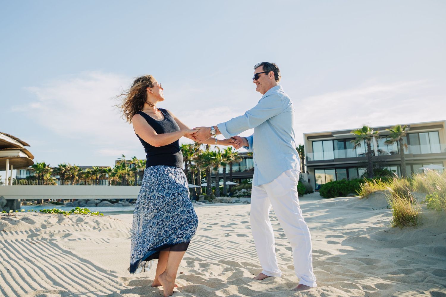 A couple dances together on a sunny beach boardwalk with palm trees and coastal homes in the background.