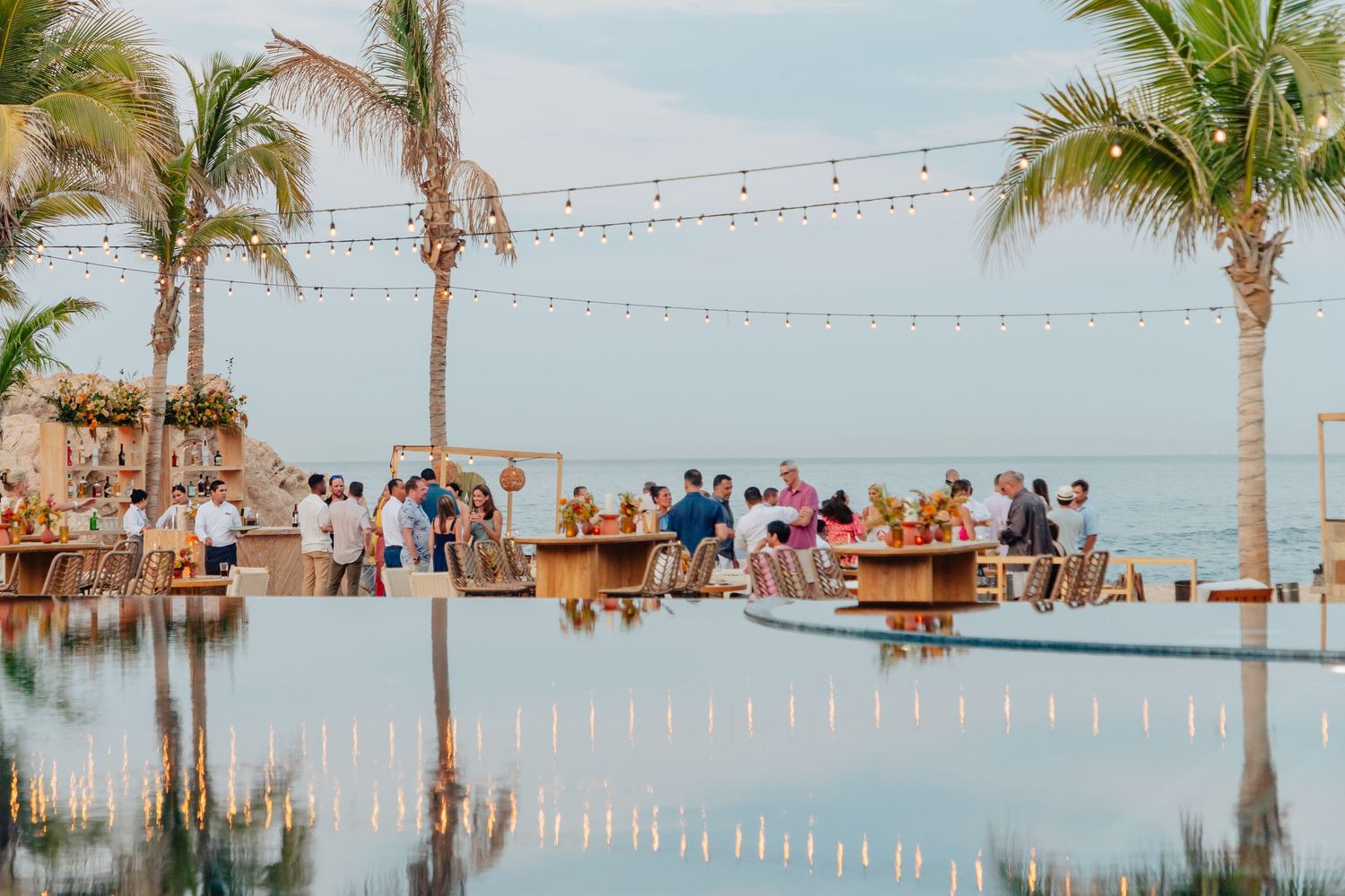 String lights illuminate a beachside venue with palm trees and ocean views at sunset.