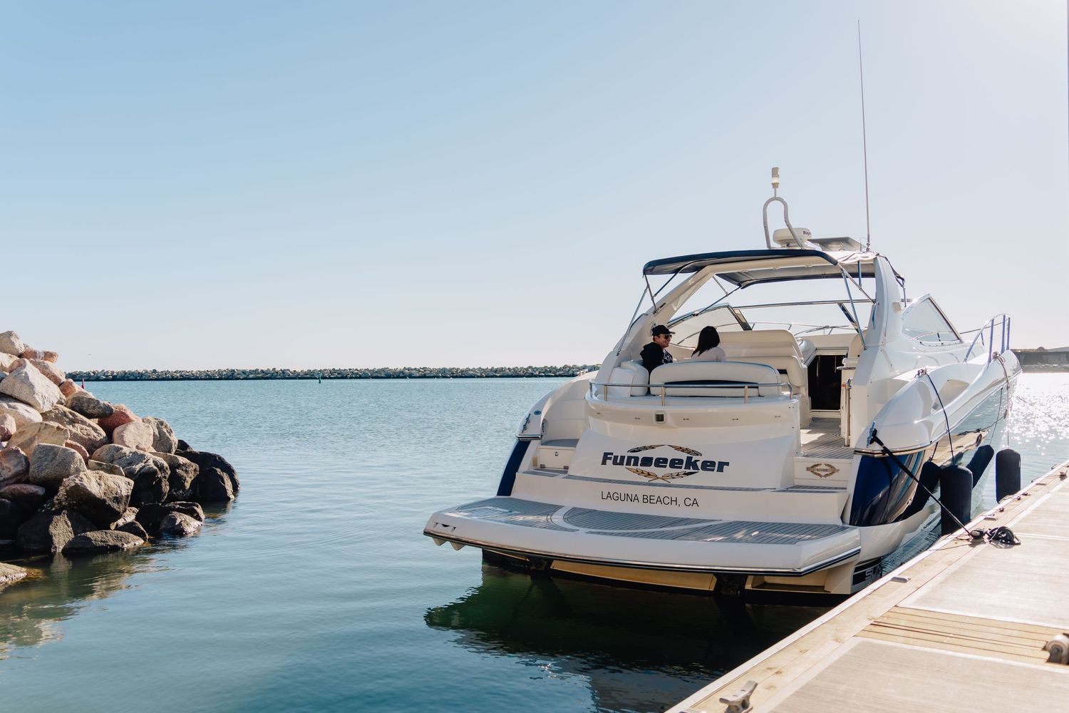 Luxury yacht moored at a marina dock next to rocky breakwater on a calm blue water day.
