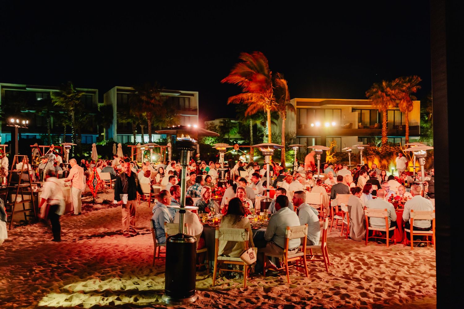 Wedding guests dance and celebrate at a beachfront reception illuminated by warm red lighting and palm trees at night.