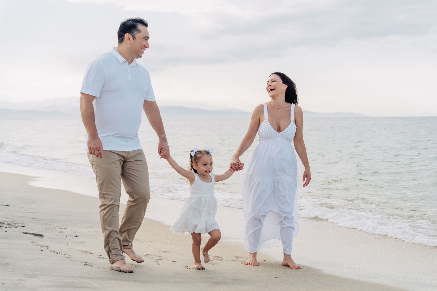 A family of three walks hand in hand along a serene beach at sunset wearing white and beige clothing.