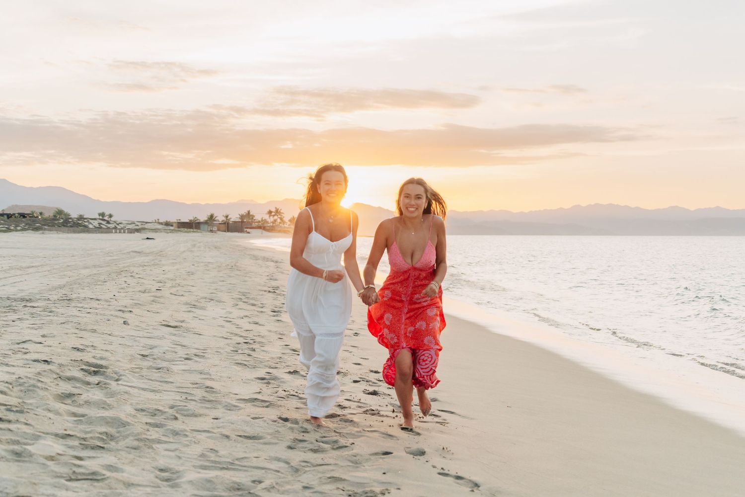 Two people in coordinating white and red outfits stroll along a sandy beach at sunset.