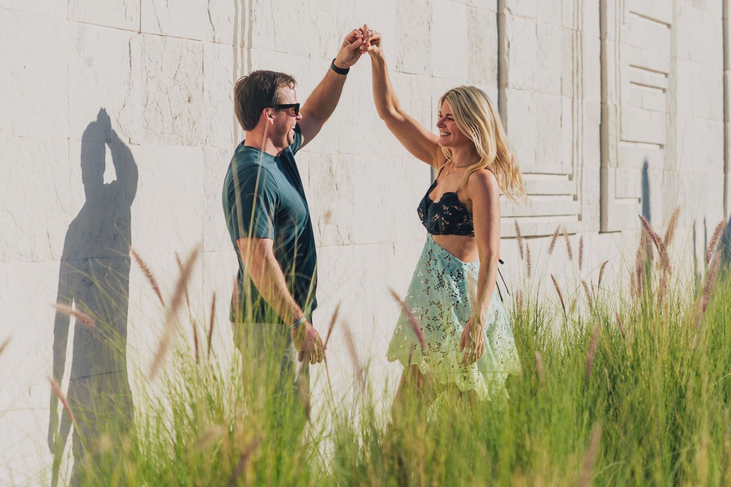 A couple holds hands while dancing in tall grass against a white wall on a summer day.