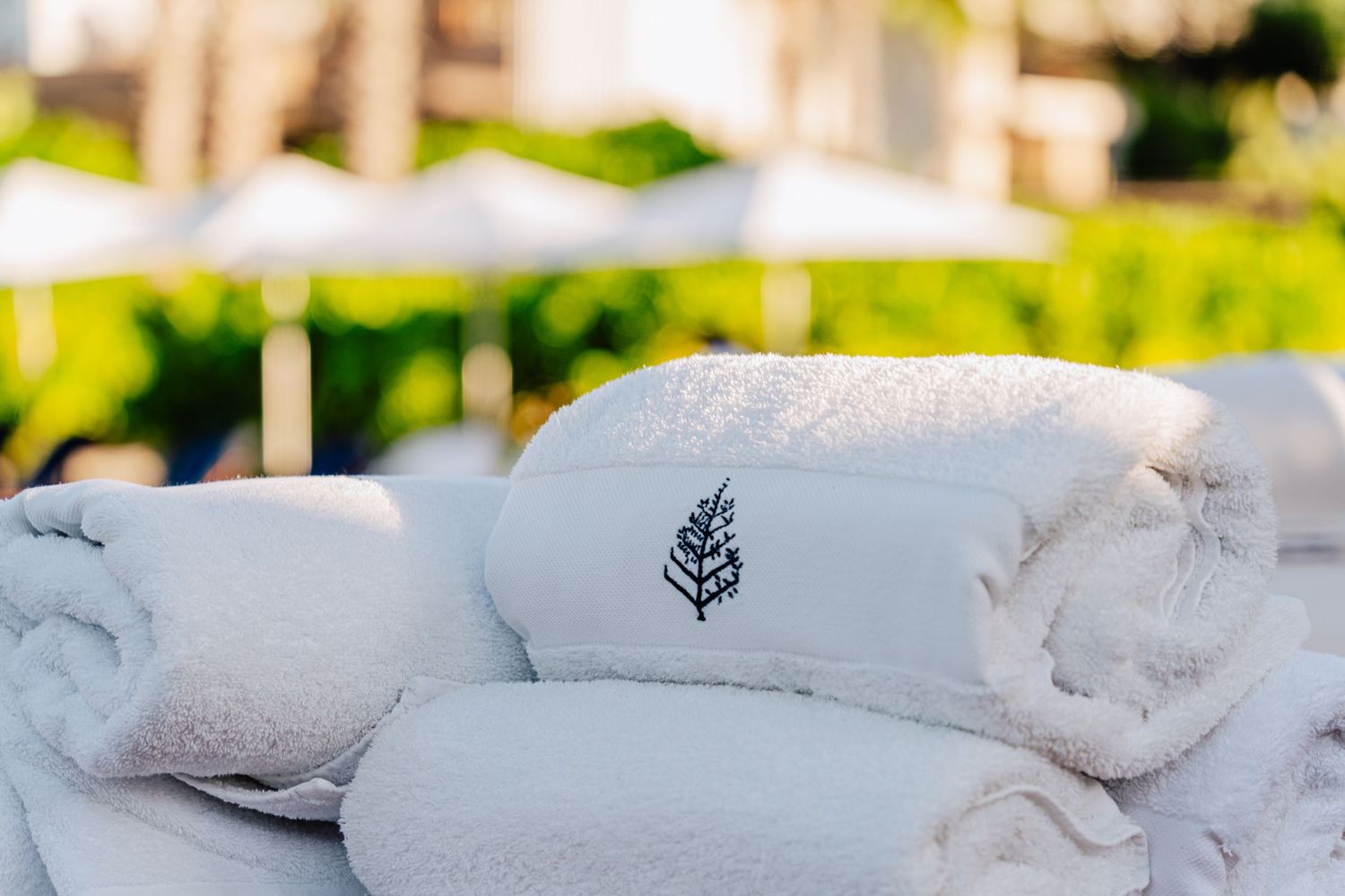 Rolled white spa towels with embroidered leaf logo stacked neatly in sunlit outdoor setting.