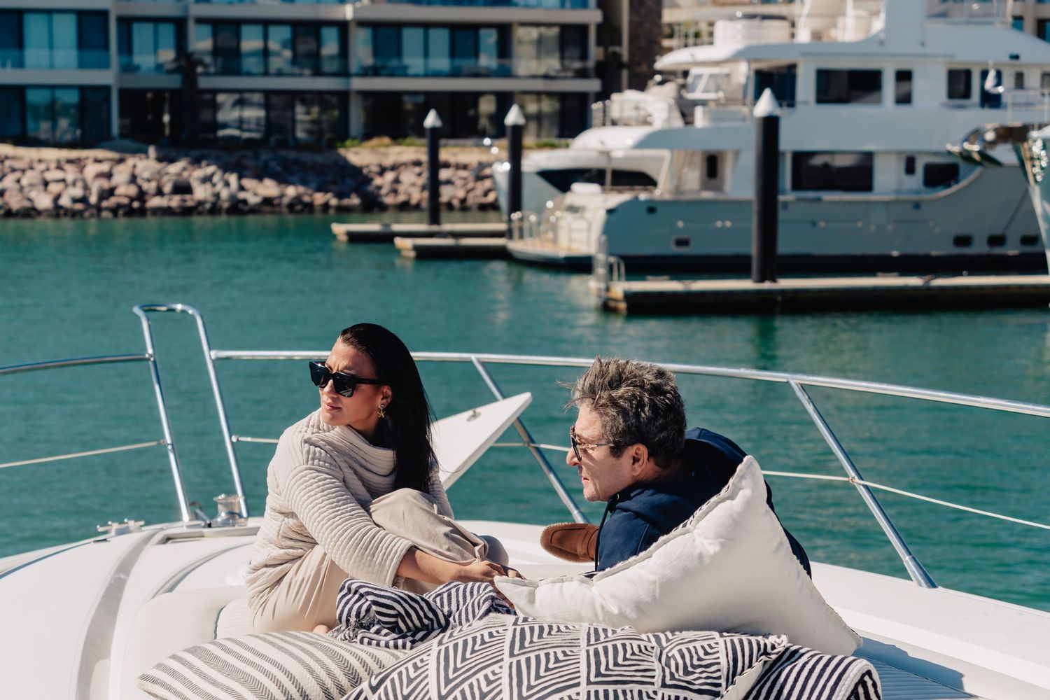 Couple relaxing on a luxury yacht deck with white cushions overlooking a marina with boats in the background.