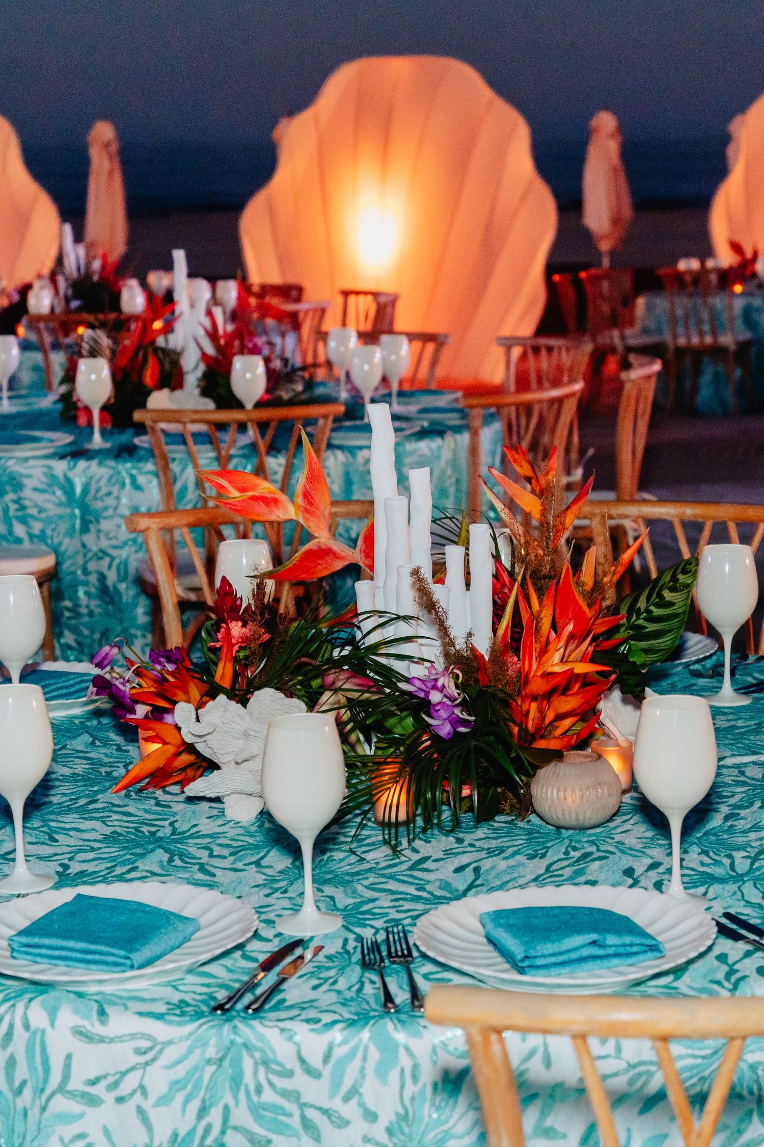 Elegant table setting with turquoise linens, white wine glasses, and tropical floral centerpieces in warm lighting.