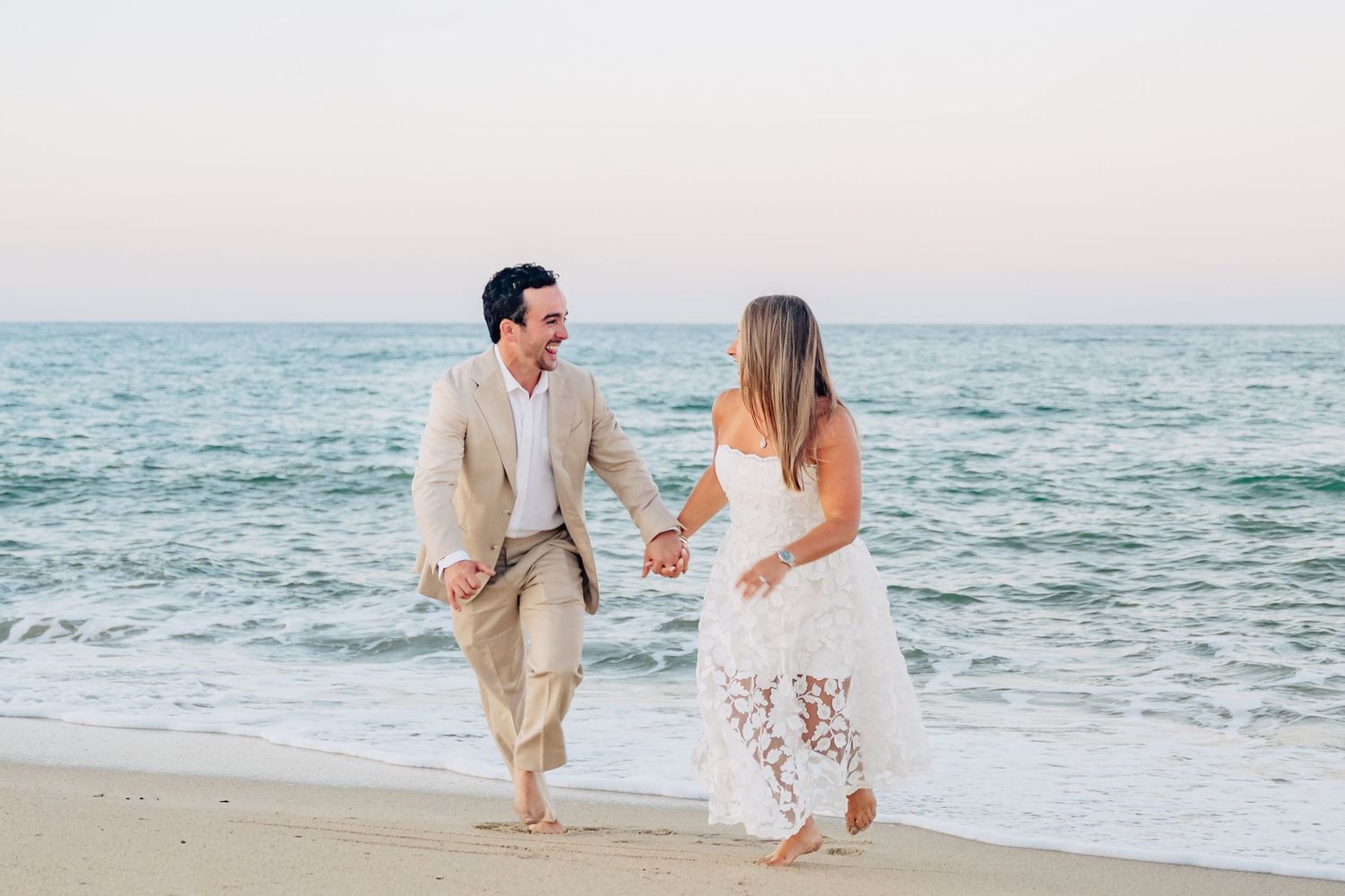 A couple in white and beige outfits walks hand in hand along a scenic beach at sunset.