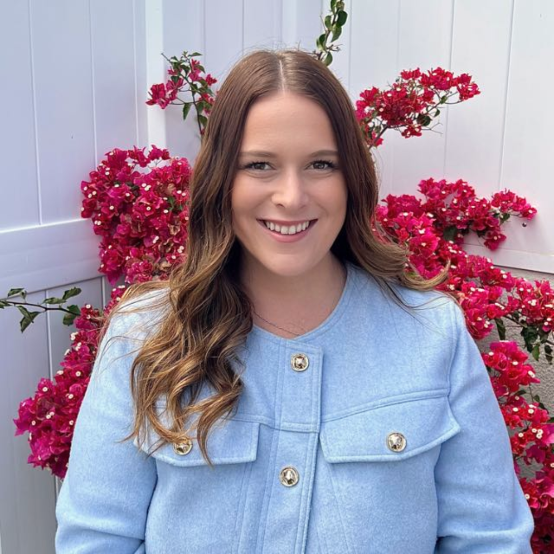 A woman in a light blue coat poses against a wall with vibrant red bougainvillea flowers.