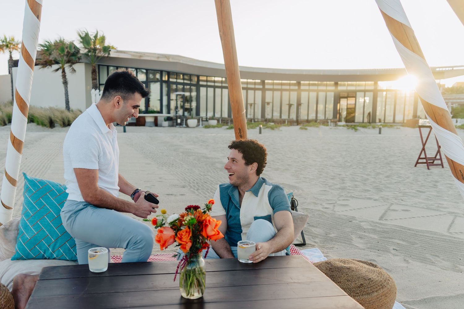A group of people sitting on beach chairs at sunset enjoying drinks and conversation on a sandy shore.