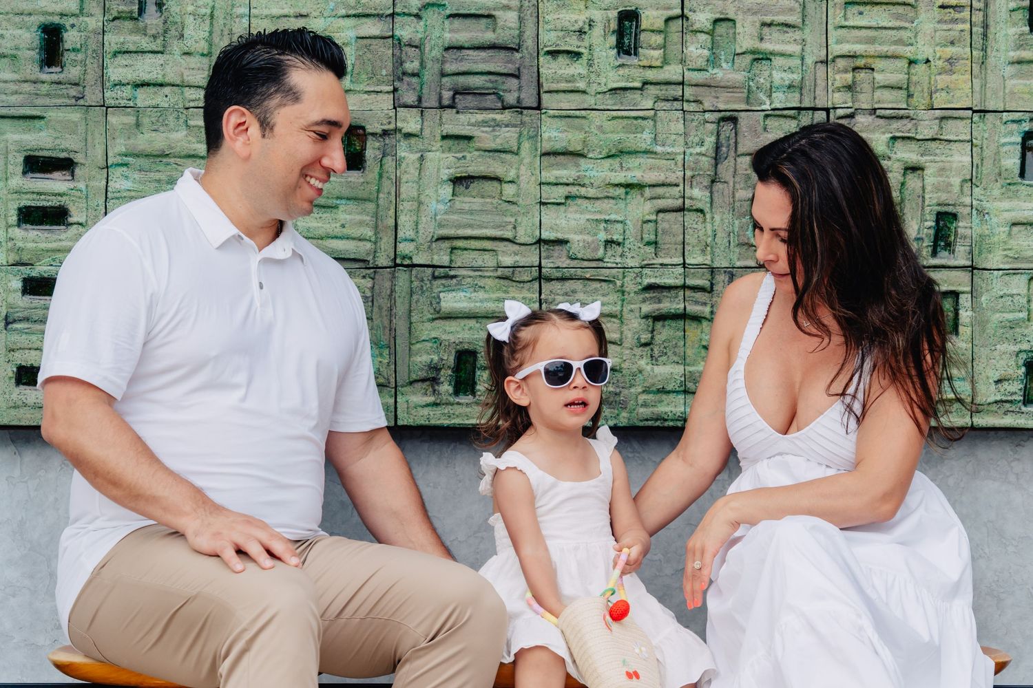 A family wearing white and khaki outfits sits together and smiles against a green textured wall background.