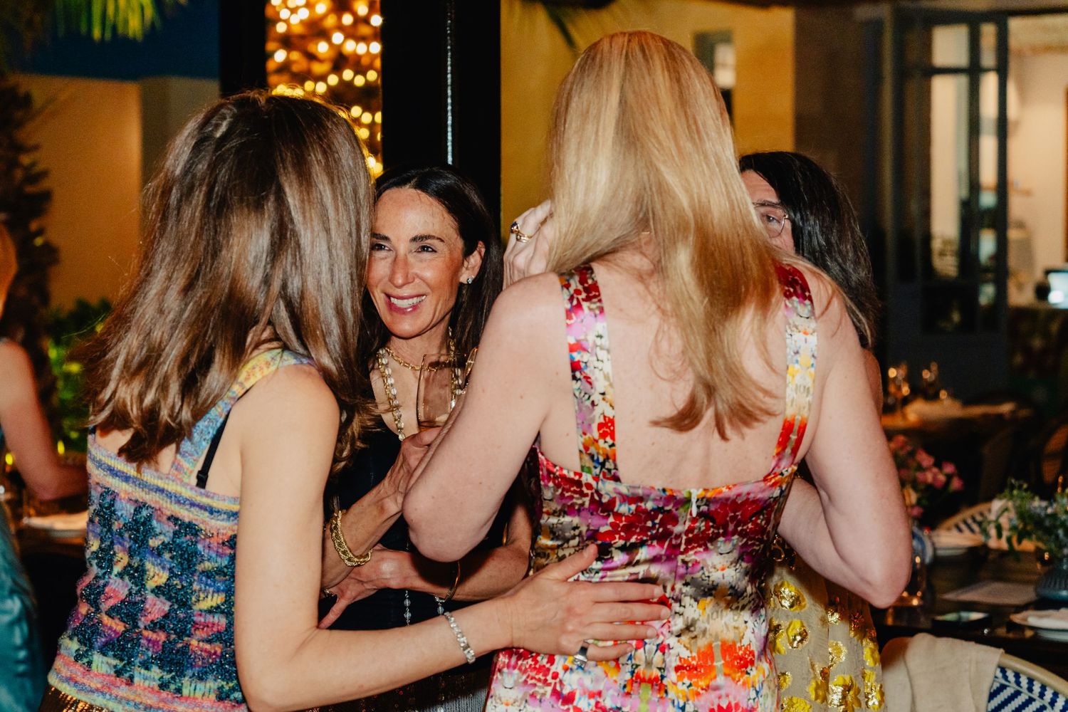 People gather at a festive social event with decorative lighting and floral arrangements in an indoor venue.