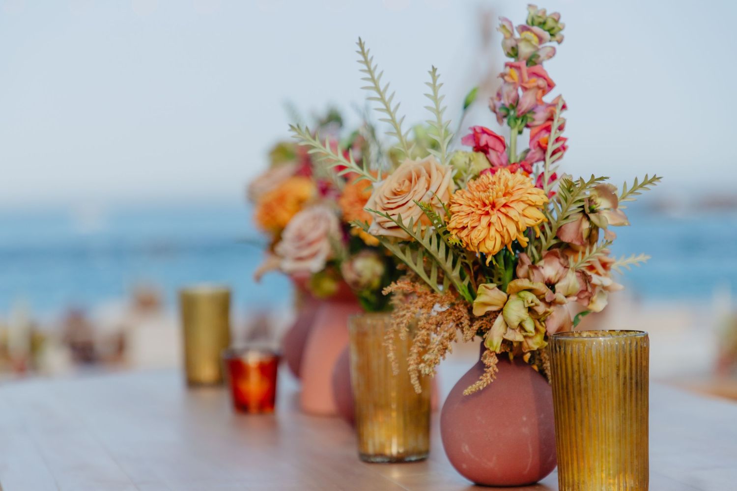 Colorful floral arrangements in amber glass vases and pink ceramic containers displayed on table with ocean view background.