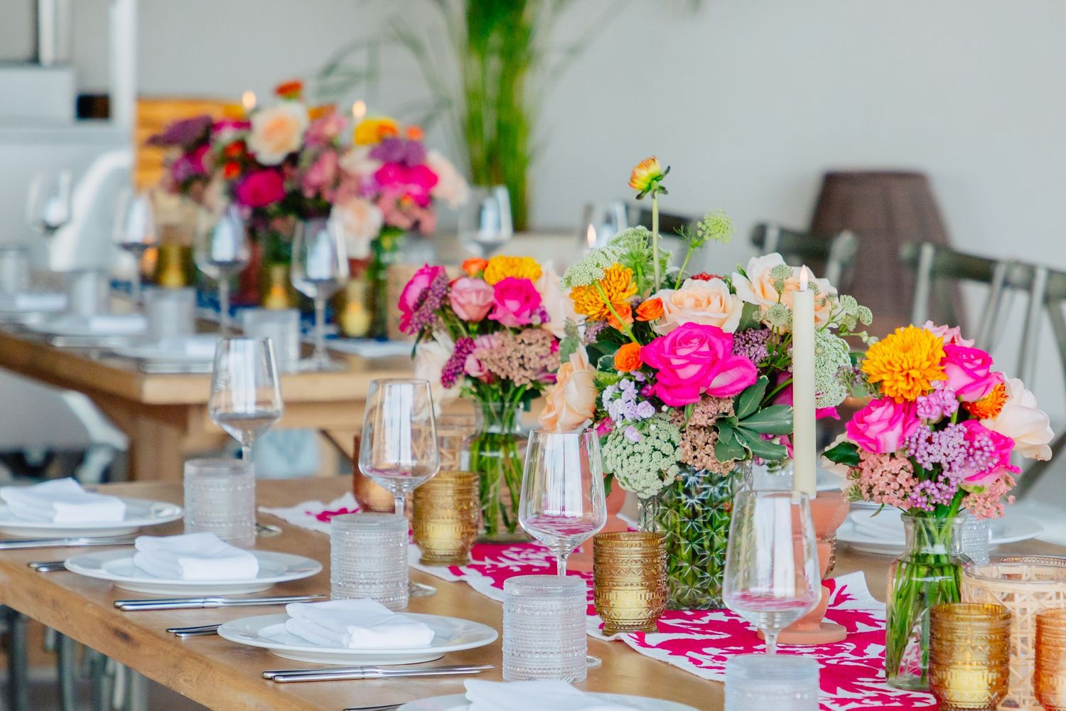 Vibrant pink and orange flower centerpieces with gold accents decorate a long wooden dining table.