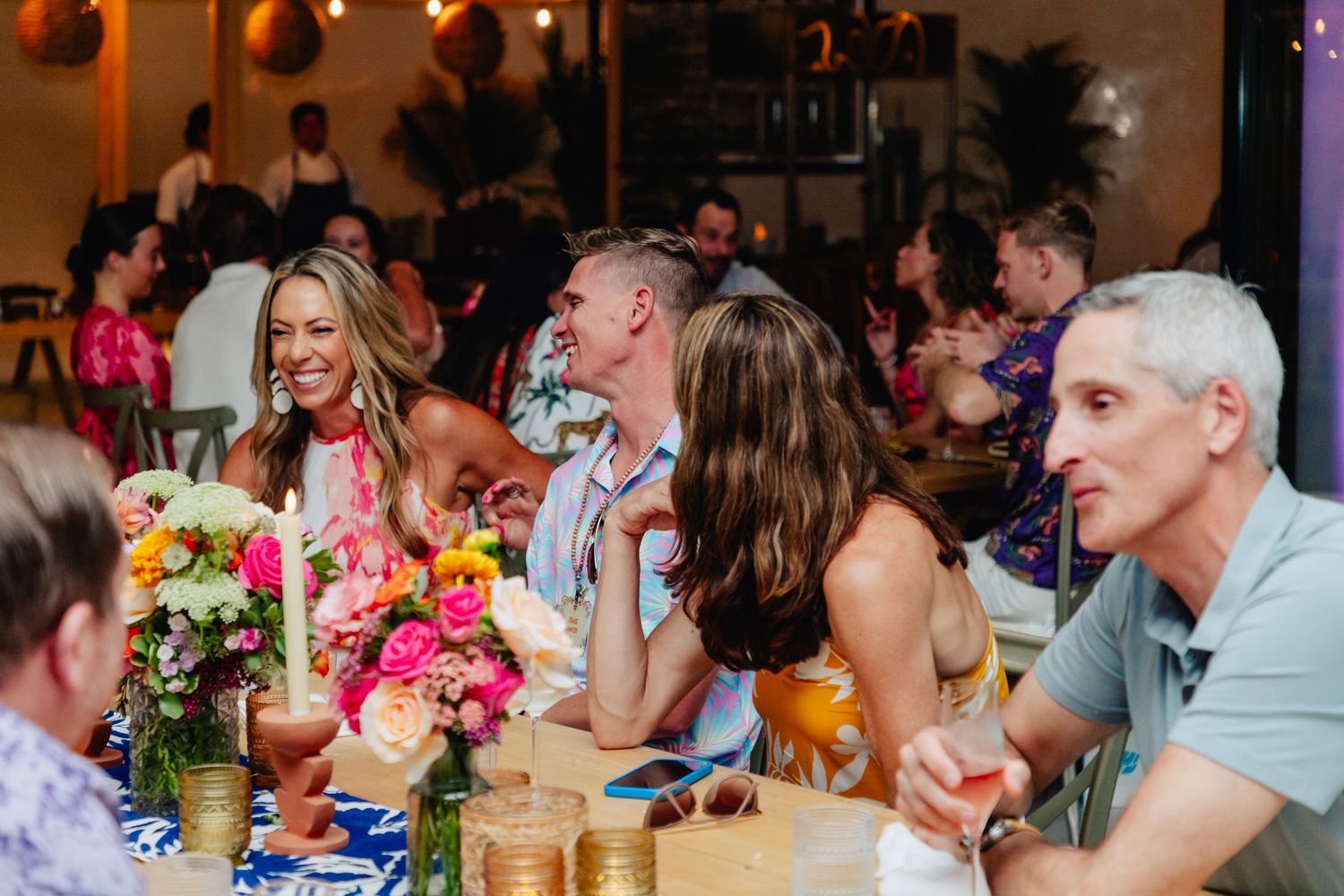 Wedding guests enjoying a lively reception dinner with colorful floral centerpieces and animated conversations at long tables.