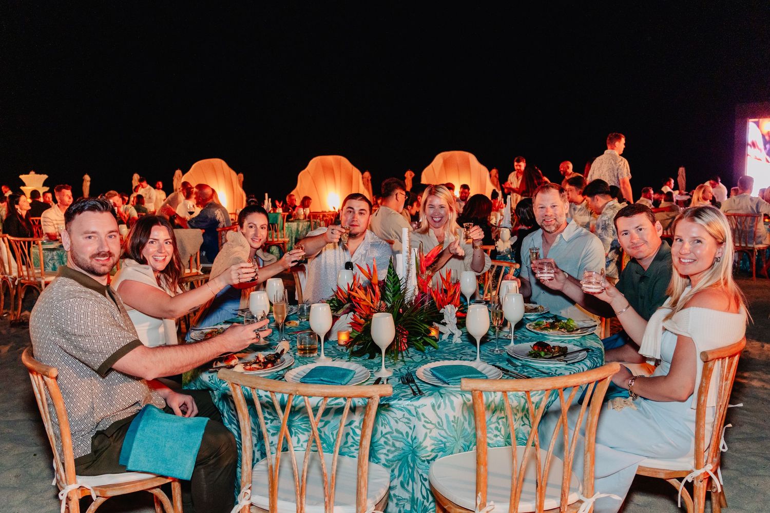 Large group of people enjoying a festive outdoor dinner party under string lights at dusk.