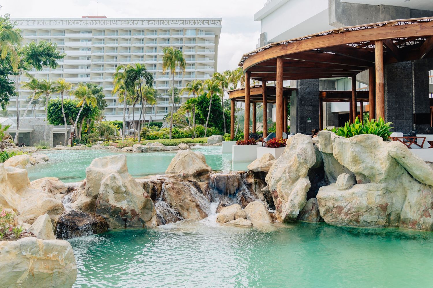 Luxury resort pool area with natural rock formations and waterfalls surrounded by palm trees.