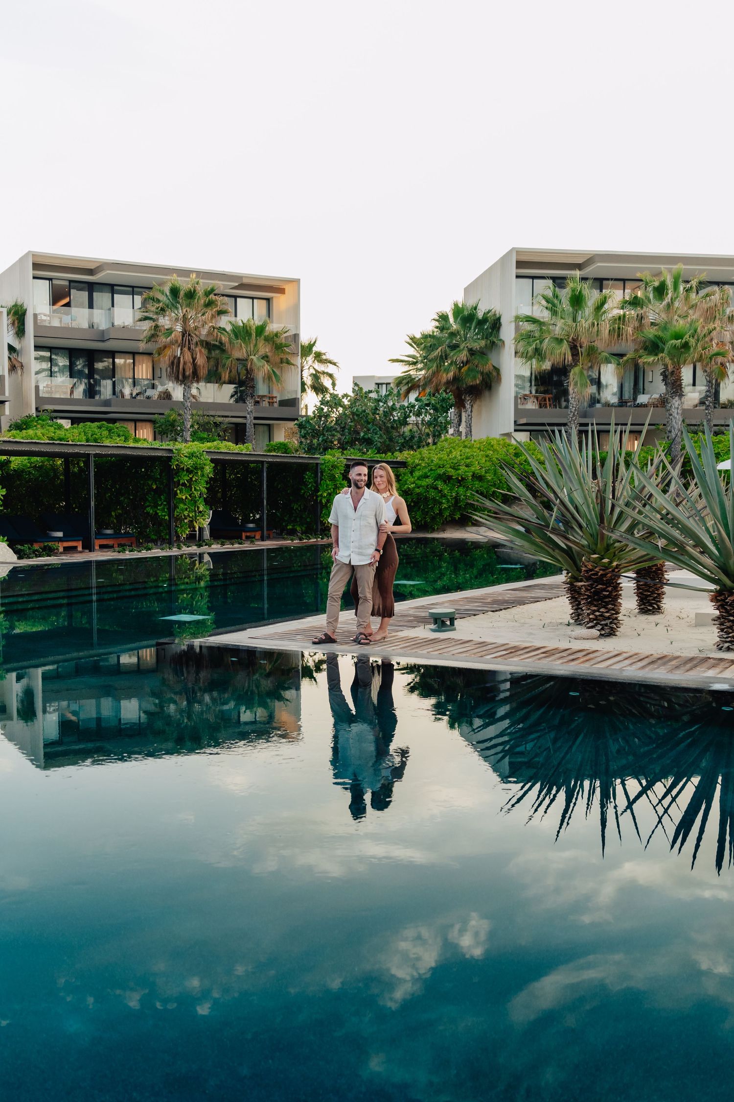 Couple in white outfits standing by a luxurious infinity pool with palm trees and modern architecture in the background.