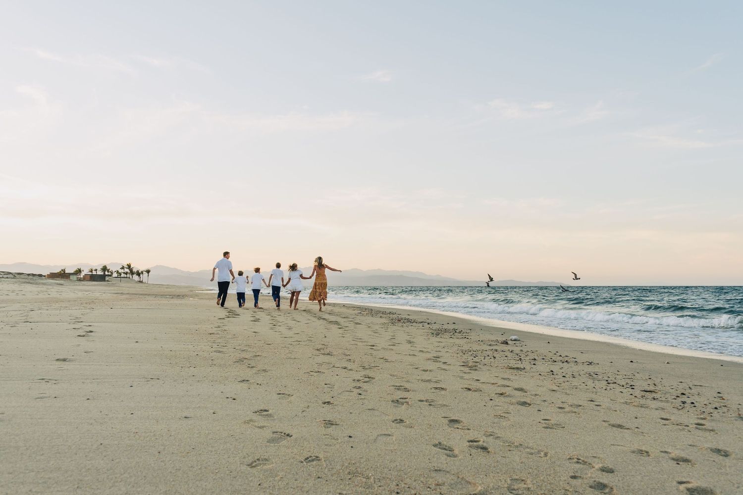 Group of people walking along a sandy beach at sunset with waves crashing on the shore and soft pastel sky colors.