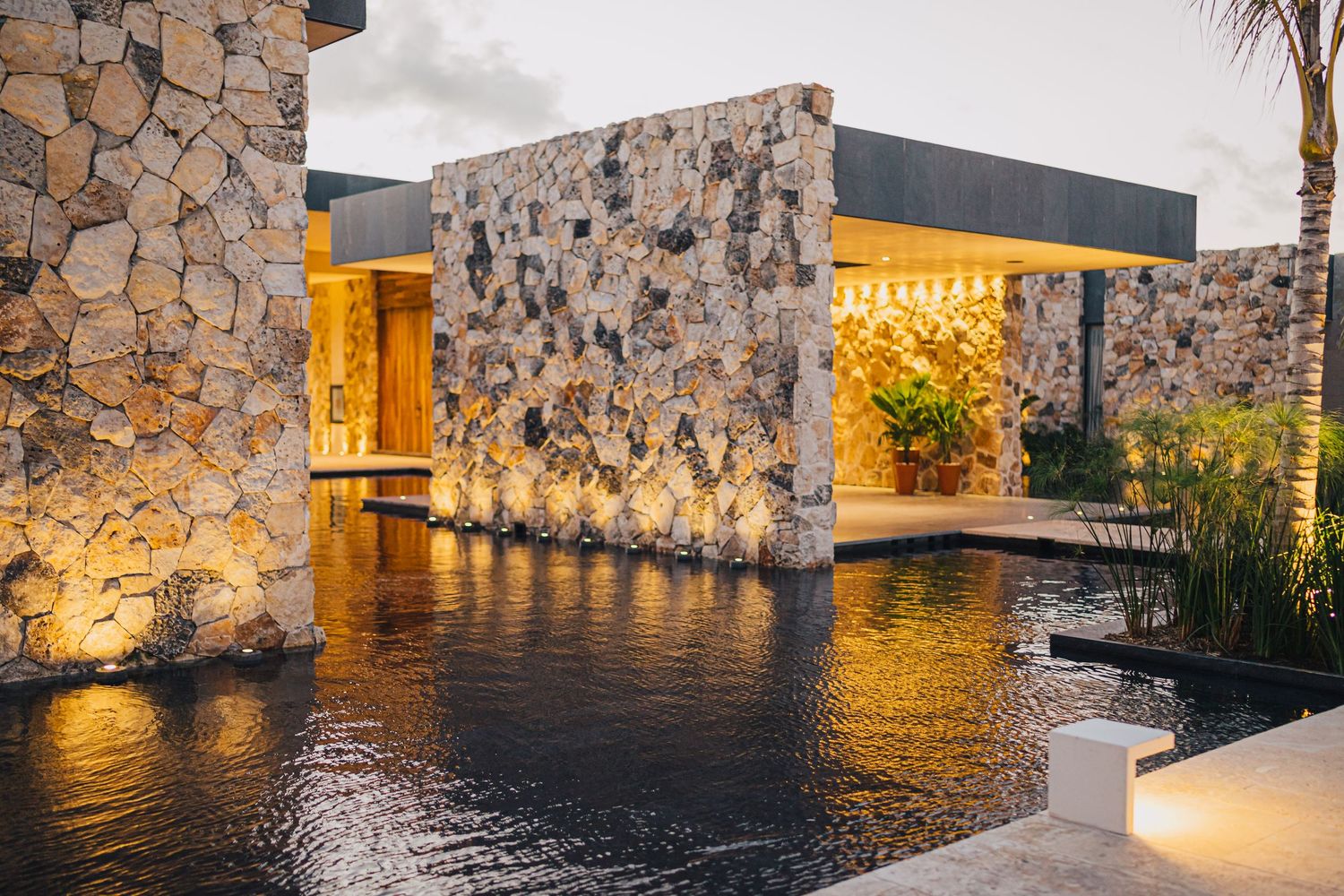Stone walls and palm trees reflect in calm water at dusk with warm lighting creating a serene atmosphere.