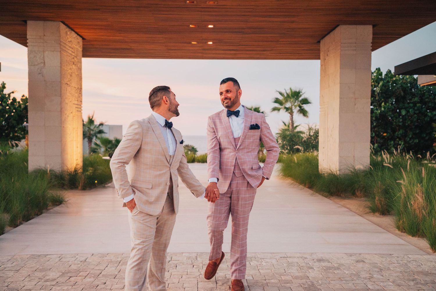 Two people in light colored suits walking and laughing under a covered outdoor walkway.
