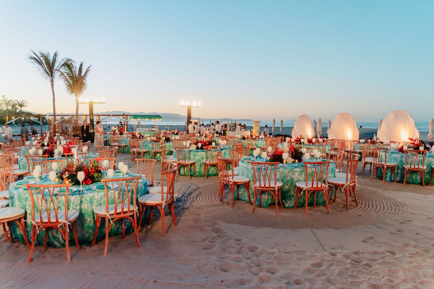 Beachfront dining tables with turquoise chairs set up along sandy shoreline at sunset during outdoor event.
