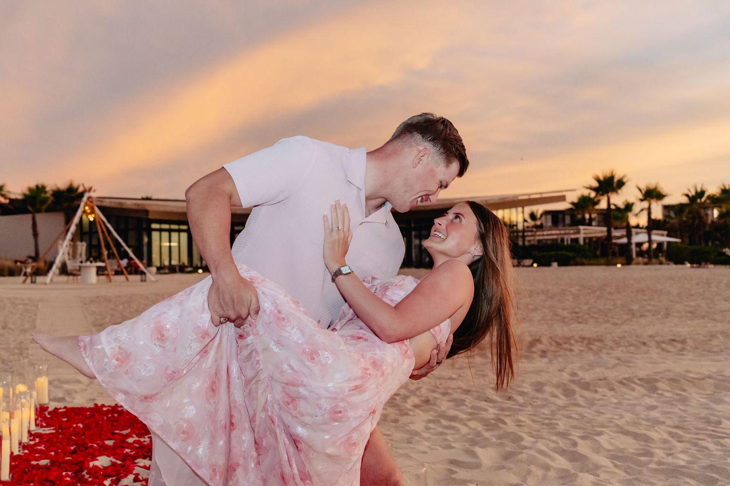 A couple in formal attire embraces on a sandy beach at sunset with palm trees in the background.
