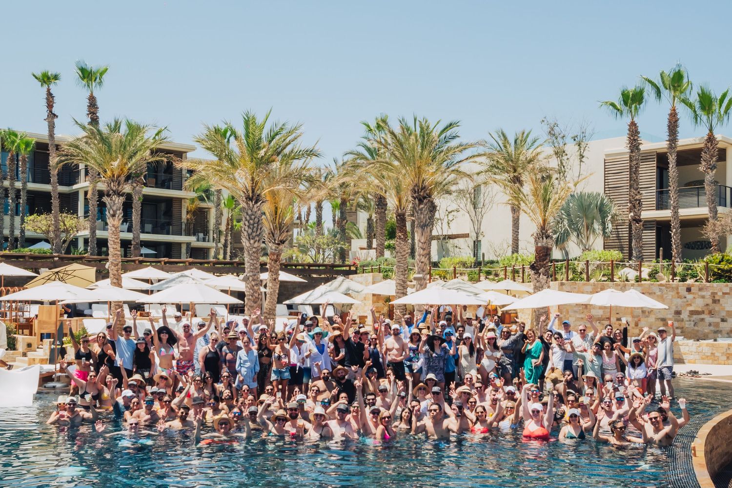 Crowded pool party scene at a resort with palm trees and white umbrellas.
