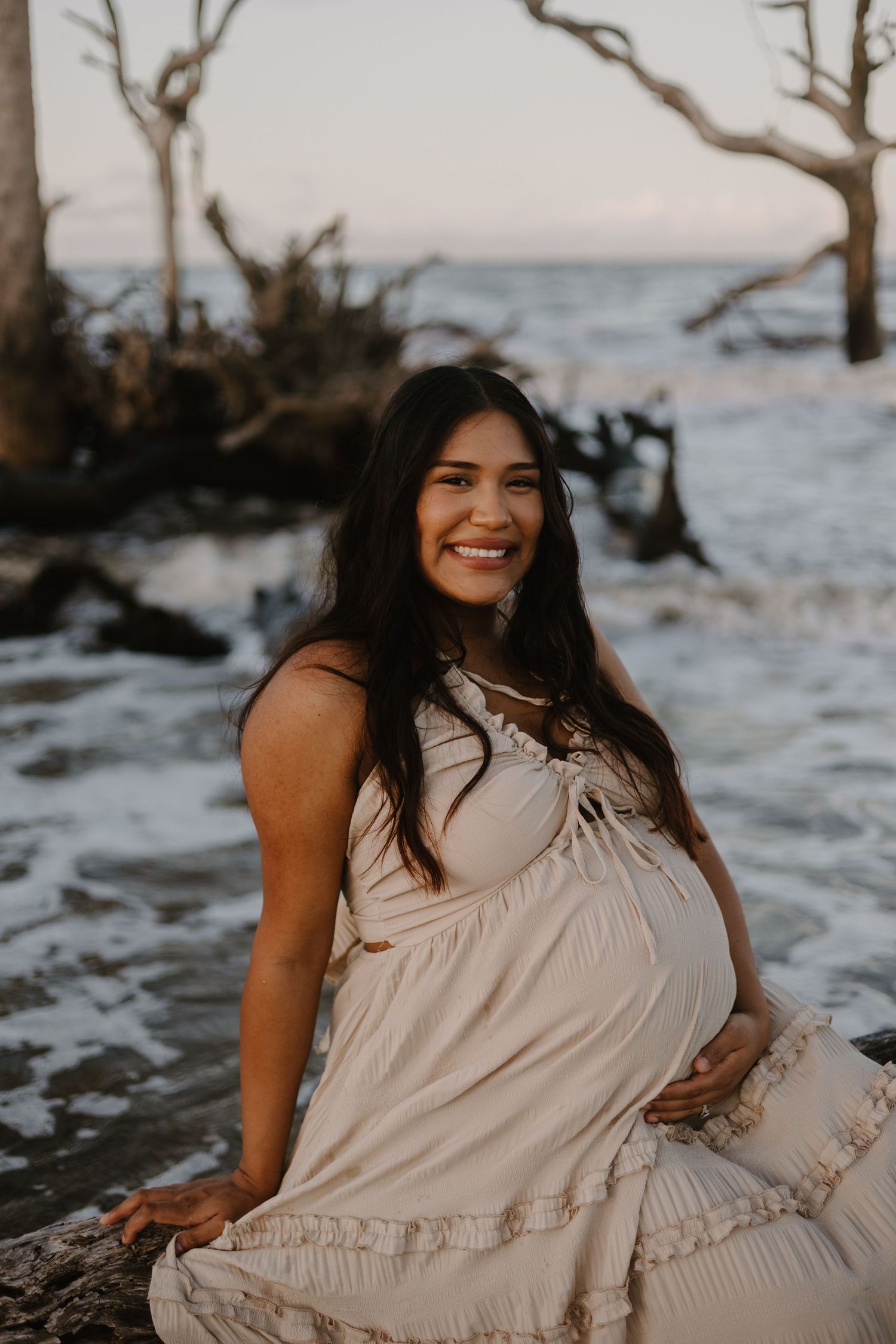Maternity photos on beach with driftwood and ocean backdrop.