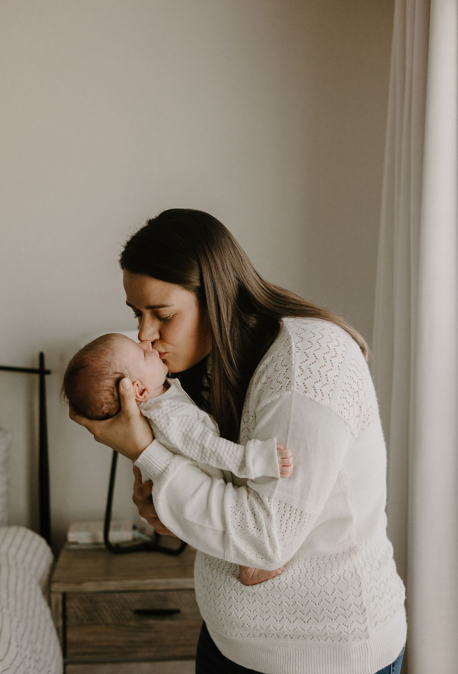 Mother in white sweater holding and kissing newborn baby indoors.