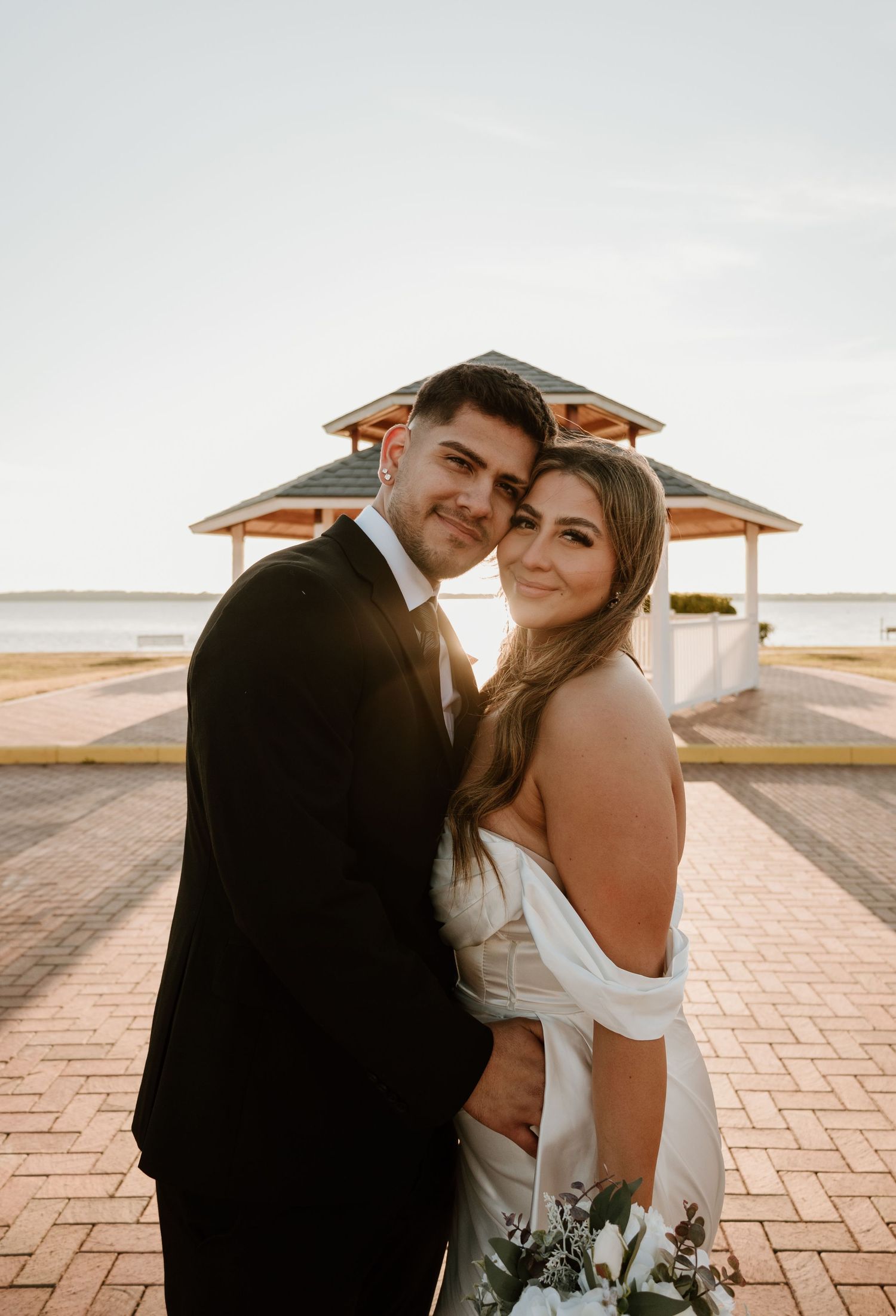 Wedding couple shares intimate moments near waterfront gazebo during golden hour.