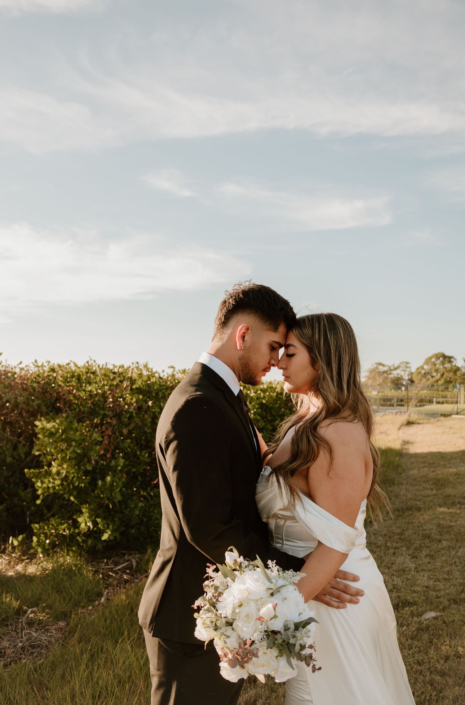 Romantic sunset wedding photos in field with couple embracing and white bouquet.