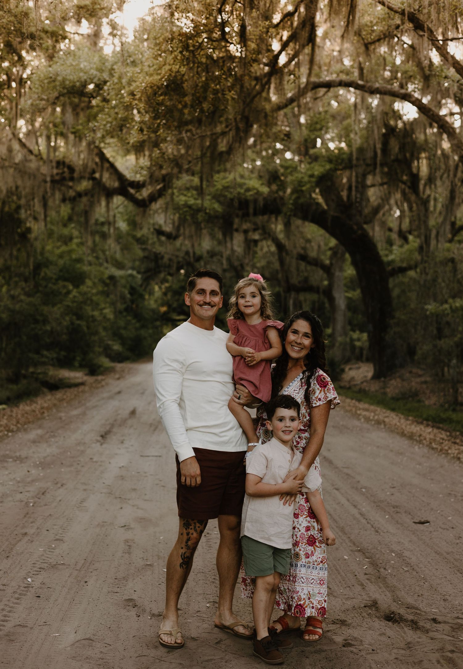 A family embraces while standing on a rustic dirt road with moss-covered trees in the background.
