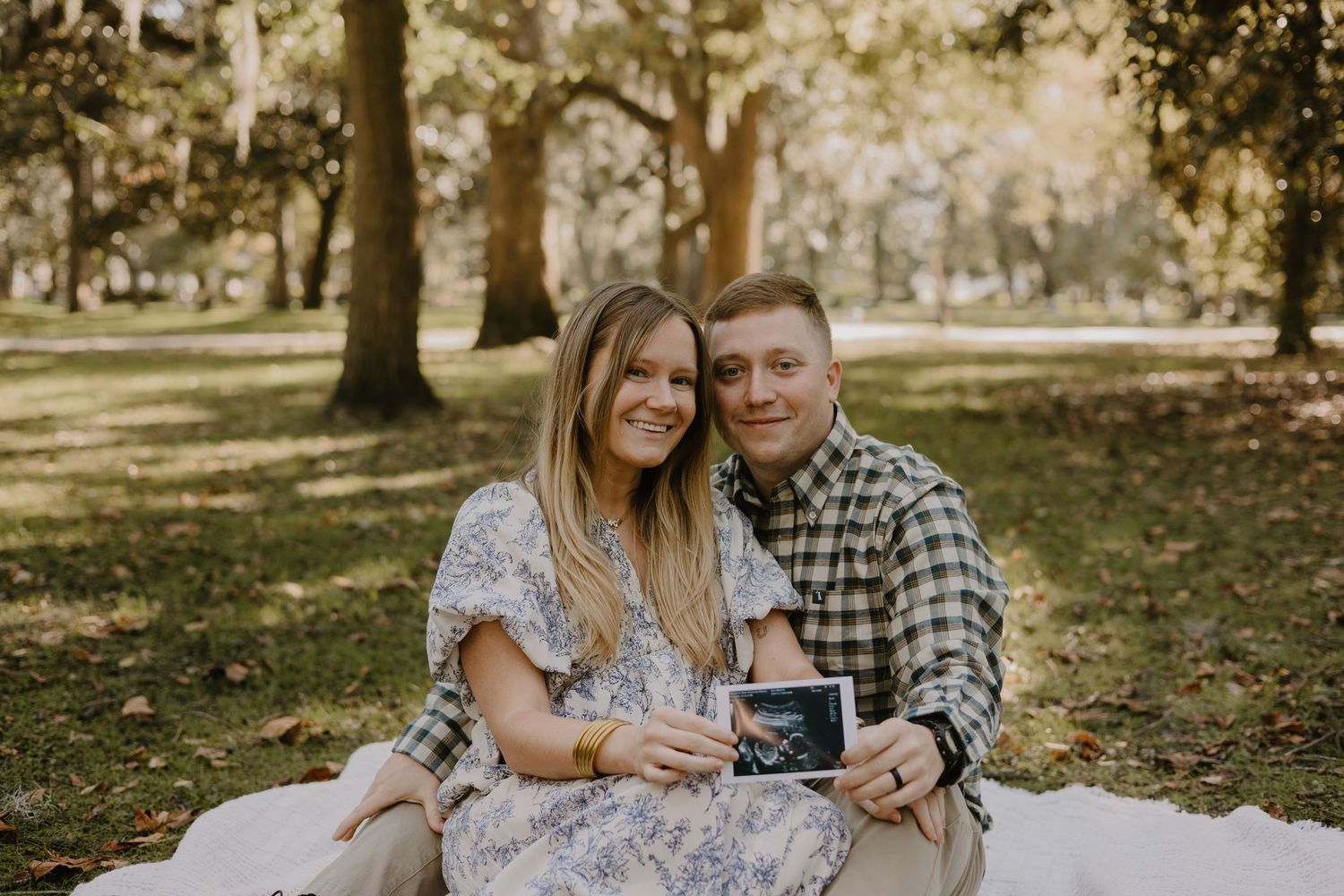 A couple shares a tender moment outdoors in a park during autumn while holding an ultrasound photo.