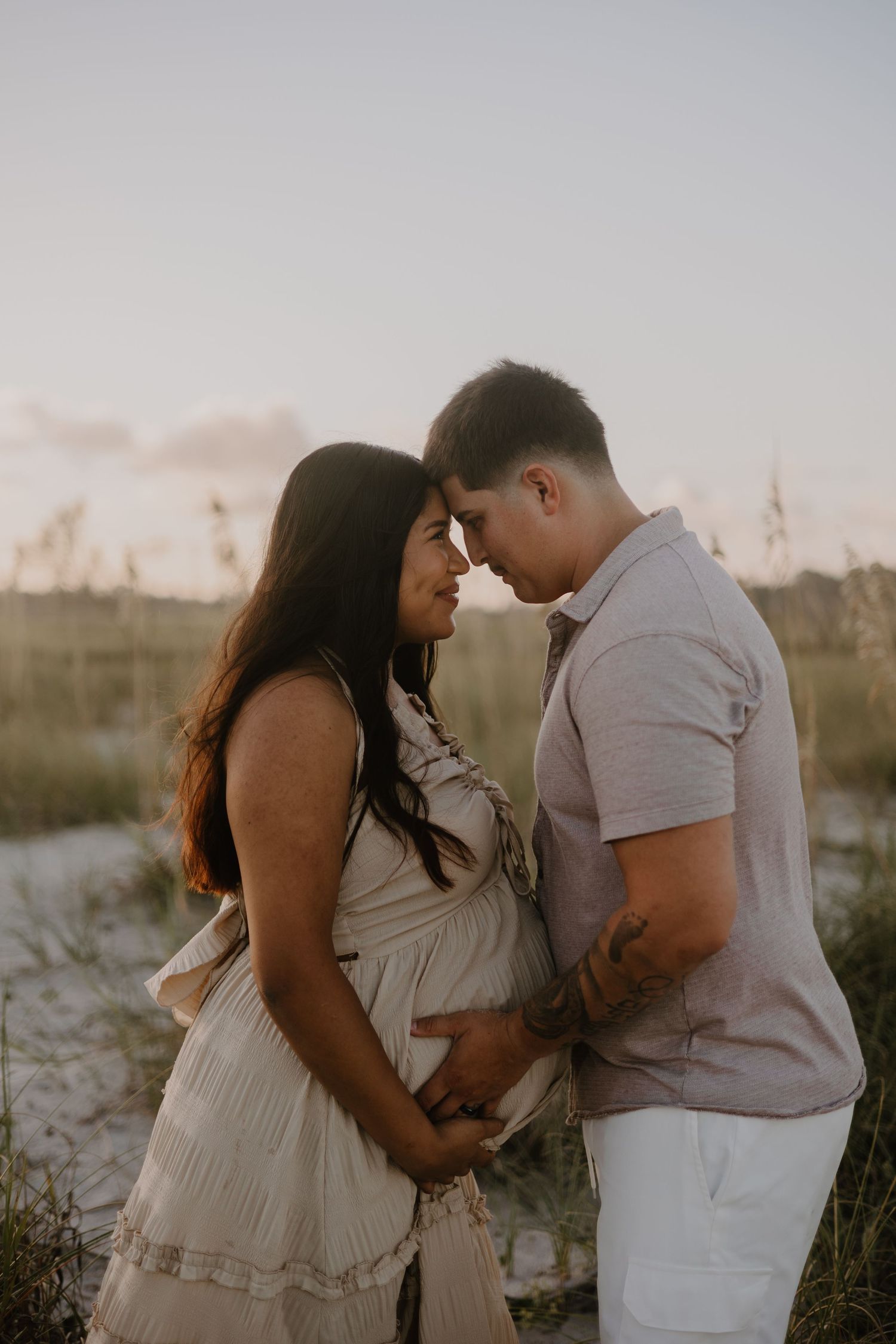 Intimate maternity photoshoot on sandy beach with couple in light neutral clothing.