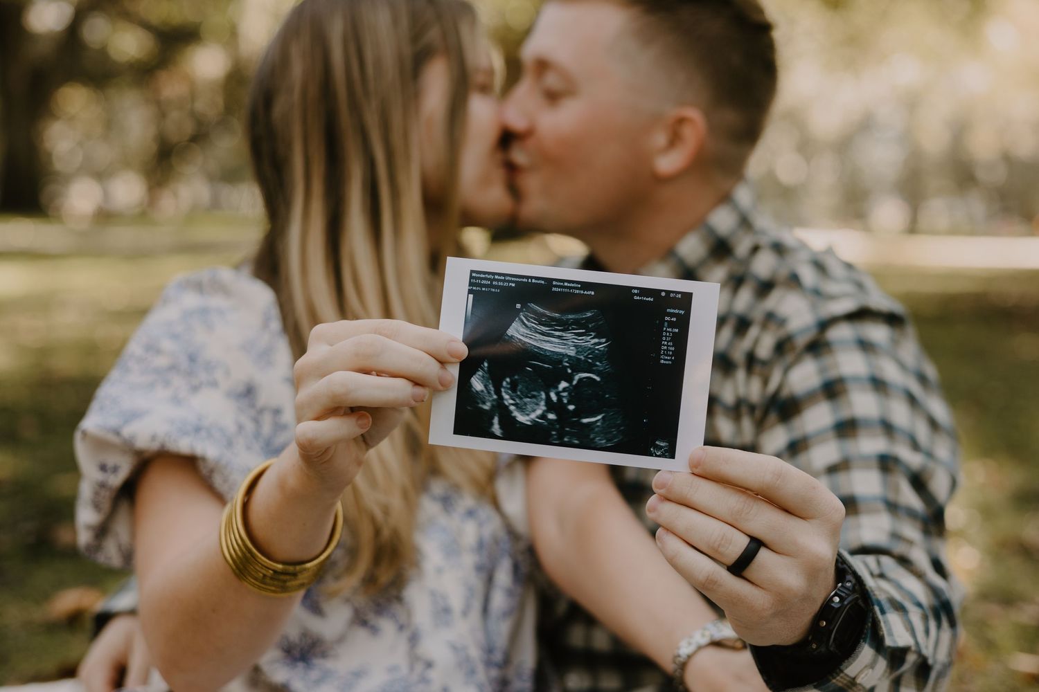 A couple holds up ultrasound photos while sharing an intimate moment outdoors.