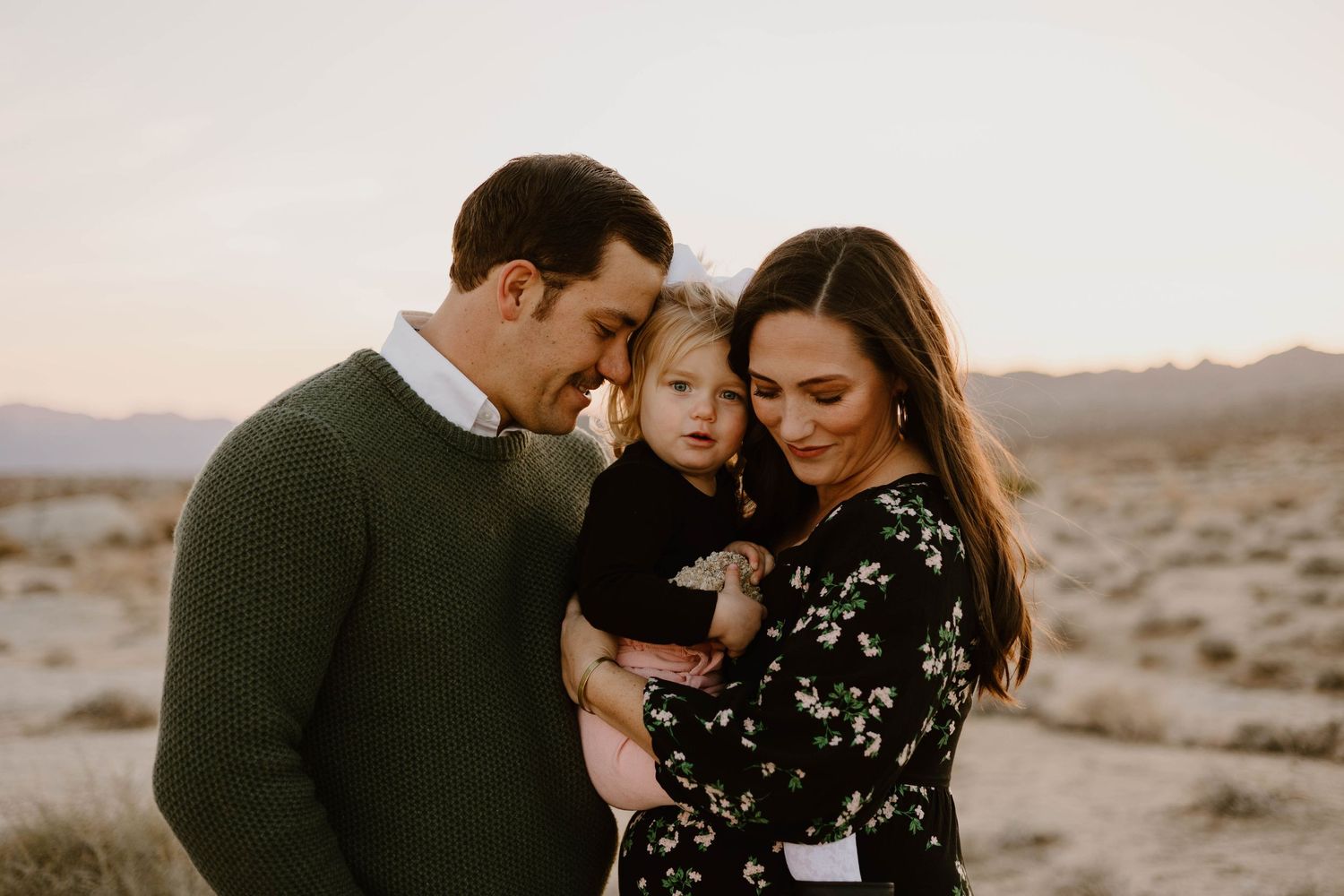 A couple and their child share intimate moments together in a desert landscape at sunset.