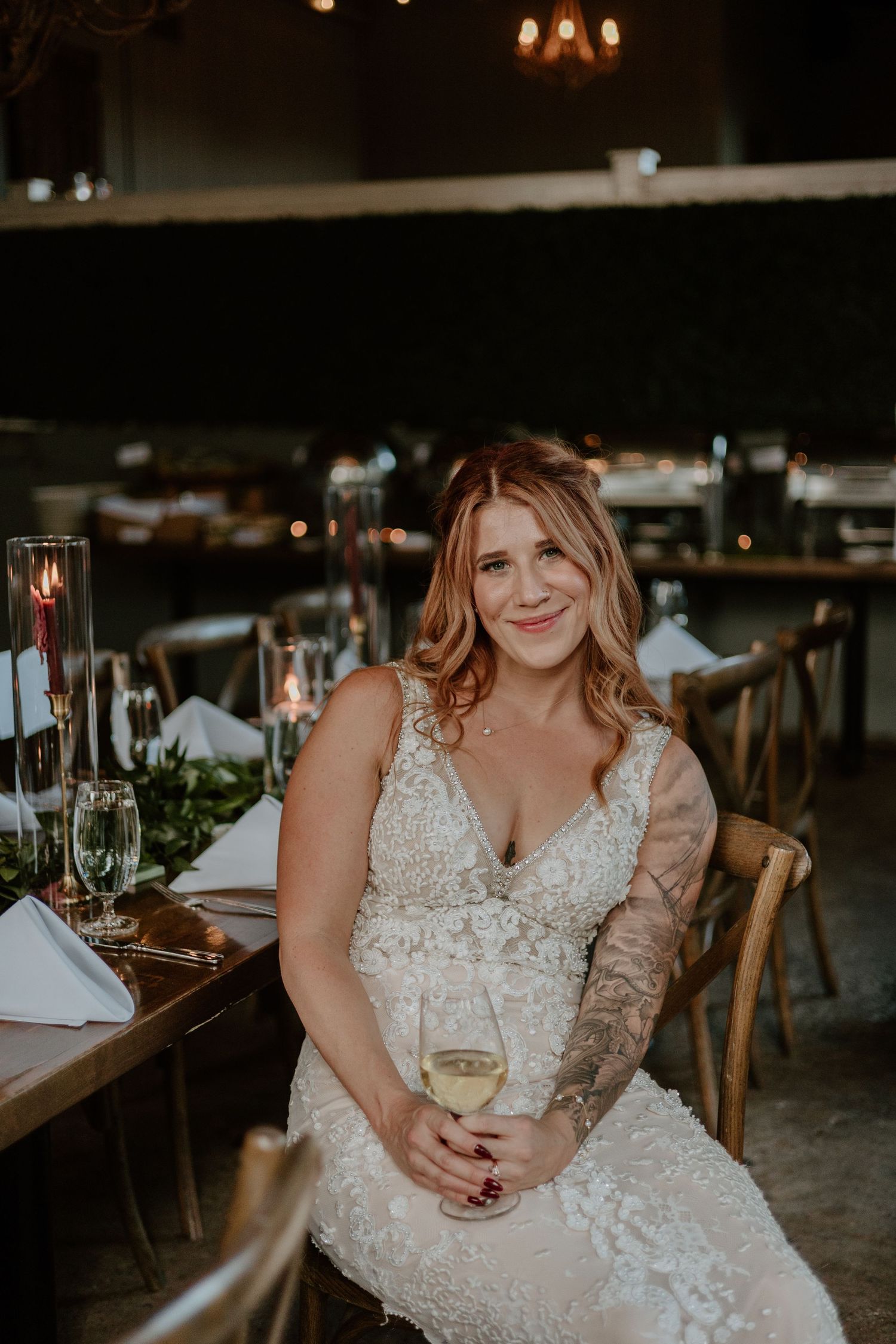 A bride in a detailed lace gown sits at a rustic reception table with glowing candlelight.