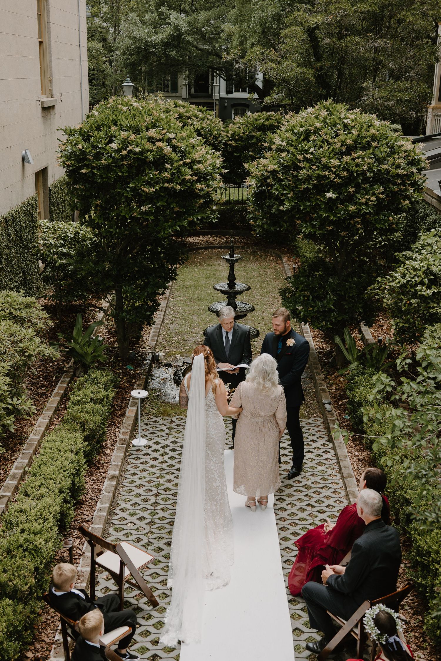 Outdoor wedding ceremony in a garden courtyard with guests seated on wooden chairs.