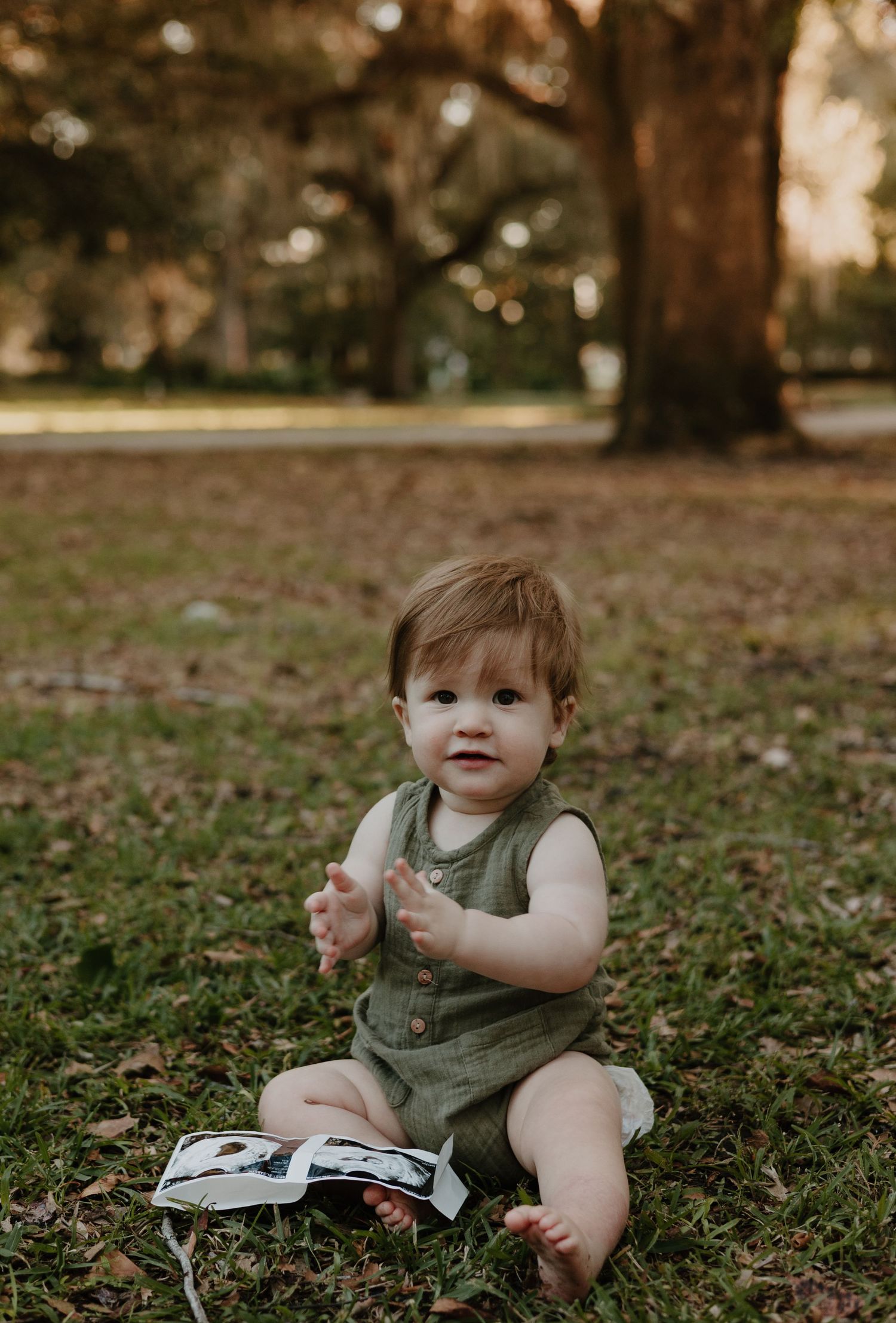 A toddler sits on grass playing with ultrasound photos during an outdoor photo session.