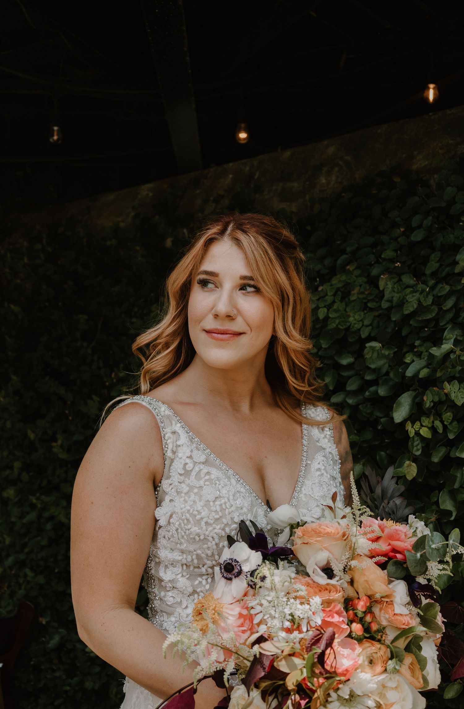 A bride in a lace dress holds a vibrant coral and peach floral bouquet against a dark garden backdrop.